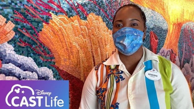 A cheerful Disney cast member sporting a blue mask beams with excitement in front of a vibrant mosaic wall, thrilled to be back welcoming guests on the Disney Fantasy cruise ship.