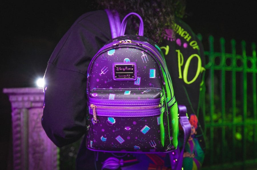 A guest shows off a vibrant, brand-new Hocus Pocus space-themed backpack by a glowing green fence at night in the theme park.