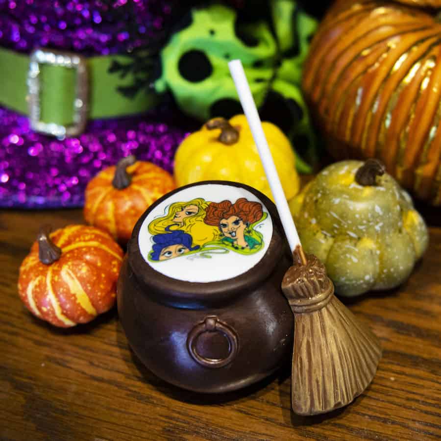 A whimsical chocolate cauldron packed with a lollipop, broomstick, and tiny pumpkins, topped by a cauldron lid showcasing the iconic Sanderson Sisters' cartoon faces—a perfect treat for Hocus Pocus fans and exclusive addition to your Disney Parks merchandise collection.