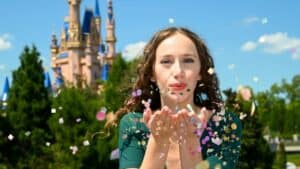 A guest at Magic Kingdom joyfully blows vibrant confetti in front of Cinderella Castle, surrounded by lush greenery under a bright blue sky, capturing the festive spirit and photo-worthy moments of Walt Disney World’s 50th Anniversary celebration.