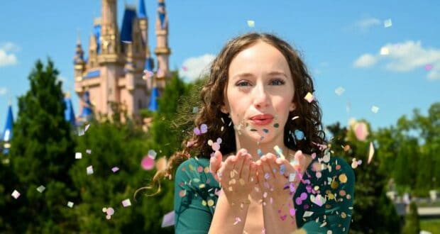 A guest at Magic Kingdom joyfully blows vibrant confetti in front of Cinderella Castle, surrounded by lush greenery under a bright blue sky, capturing the festive spirit and photo-worthy moments of Walt Disney World’s 50th Anniversary celebration.