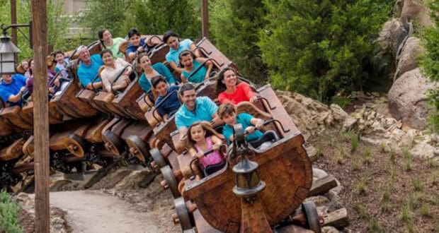 A group of people of various ages ride a wooden-themed roller coaster through a lush, green area, smiling and raising their hands in excitement—one of the top Magic Kingdom ride strategies for maximizing fun.