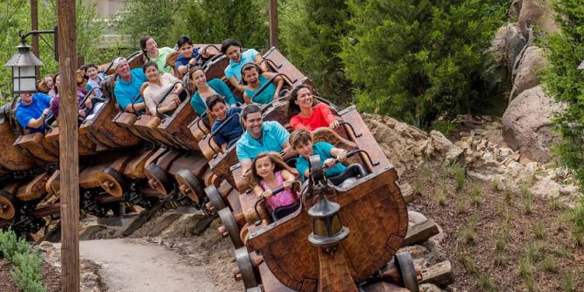A group of people of various ages ride a wooden-themed roller coaster through a lush, green area, smiling and raising their hands in excitement—one of the top Magic Kingdom ride strategies for maximizing fun.