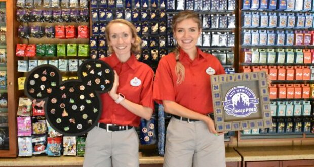 Cast Members in bright red shirts hold vibrant Disney pin displays, standing in front of a colorful pin trading wall at Walt Disney World, excited to showcase the latest ways to trade pins and help Guests get the most out of their pin trading adventure.