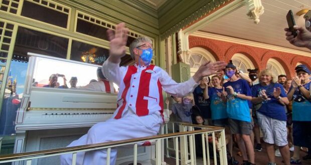A Main Street U.S.A. pianist, dressed in a signature red and white vest, entertains guests with lively tunes on a white piano during the 50th anniversary celebration as fans cheer and capture the moment with photos.