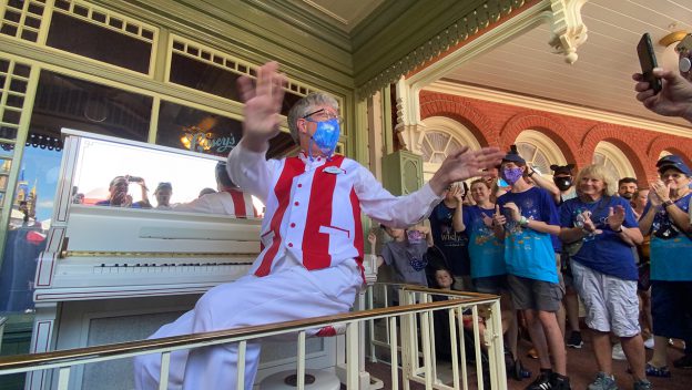 A Main Street U.S.A. pianist, dressed in a signature red and white vest, entertains guests with lively tunes on a white piano during the 50th anniversary celebration as fans cheer and capture the moment with photos.