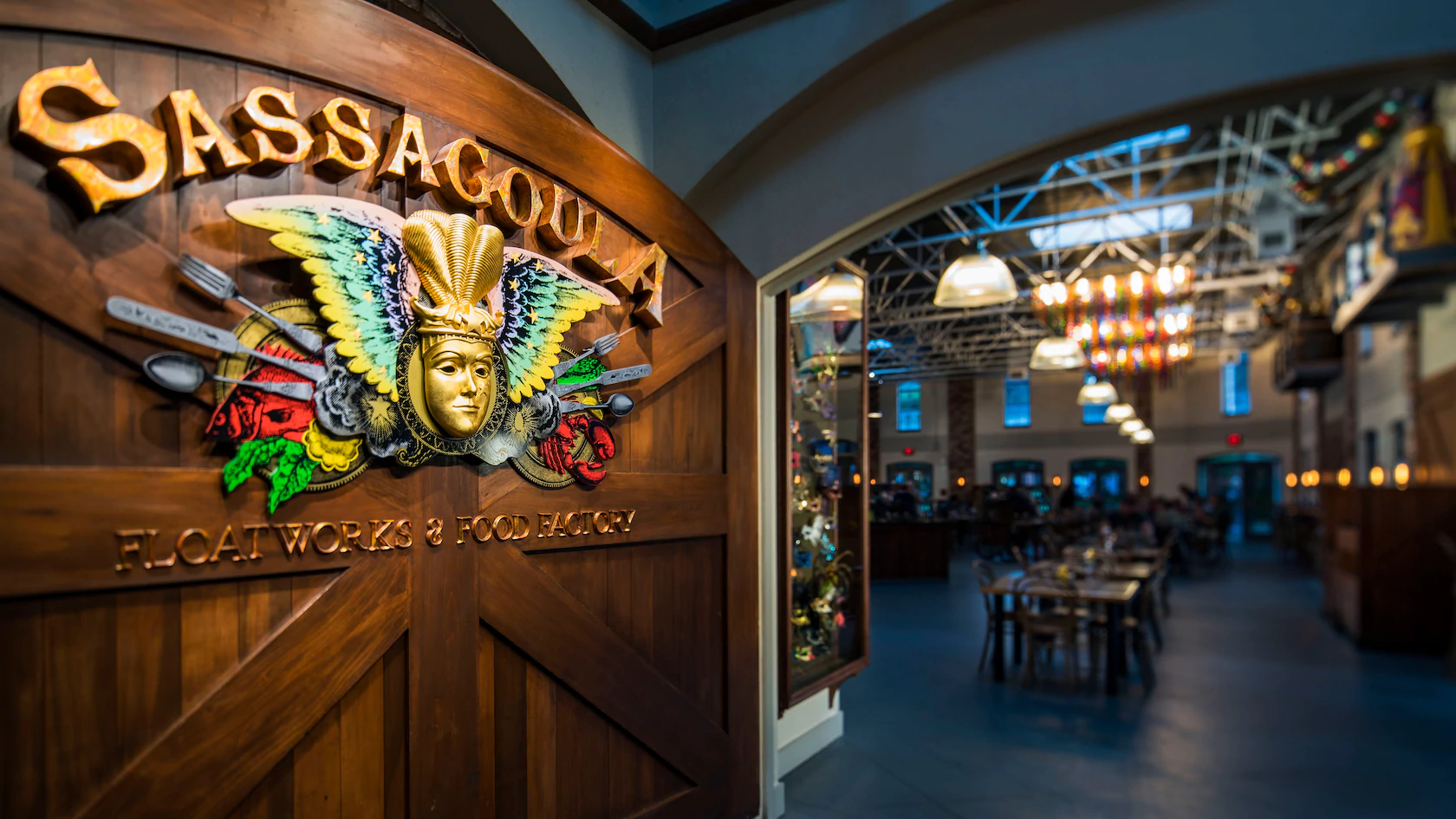 Entrance to Sassagoula Floatworks & Food Factory at Disney’s Port Orleans French Quarter, featuring a vibrant festive mask above the sign and a peek into the energetic restaurant interior where guests can enjoy famous Mickey-shaped Beignets.