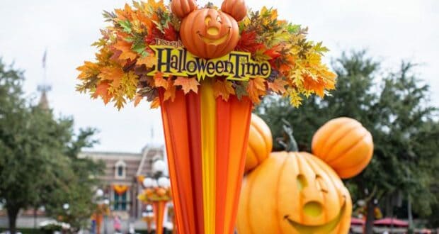 The iconic HalloweenTime sign topped with a Mickey pumpkin welcomes guests at Disneyland, surrounded by vibrant autumn leaves and a giant Mickey pumpkin glowing in the background—perfectly capturing the West Coast park’s festive Halloween atmosphere.