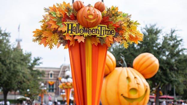 The iconic HalloweenTime sign topped with a Mickey pumpkin welcomes guests at Disneyland, surrounded by vibrant autumn leaves and a giant Mickey pumpkin glowing in the background—perfectly capturing the West Coast park’s festive Halloween atmosphere.