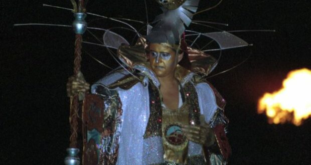 A dazzling Tapestry of Nations performer wears a shimmering gold and silver costume and ornate helmet, grasping a staff as flames blaze in the background, capturing the grandeur and excitement of EPCOT’s iconic parade.