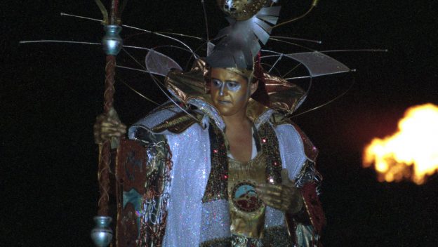 A dazzling Tapestry of Nations performer wears a shimmering gold and silver costume and ornate helmet, grasping a staff as flames blaze in the background, capturing the grandeur and excitement of EPCOT’s iconic parade.