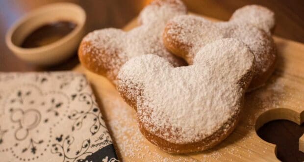Step into Disney magic with three Mickey-shaped beignets dusted in powdered sugar, served on a wooden board alongside a bowl of rich chocolate sauce—bringing the iconic Port Orleans French Quarter treat to your theme park adventure.