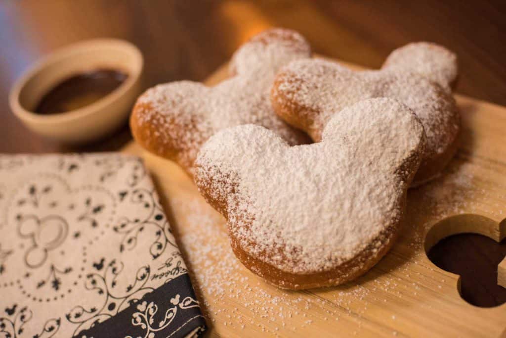 Step into Disney magic with three Mickey-shaped beignets dusted in powdered sugar, served on a wooden board alongside a bowl of rich chocolate sauce—bringing the iconic Port Orleans French Quarter treat to your theme park adventure.