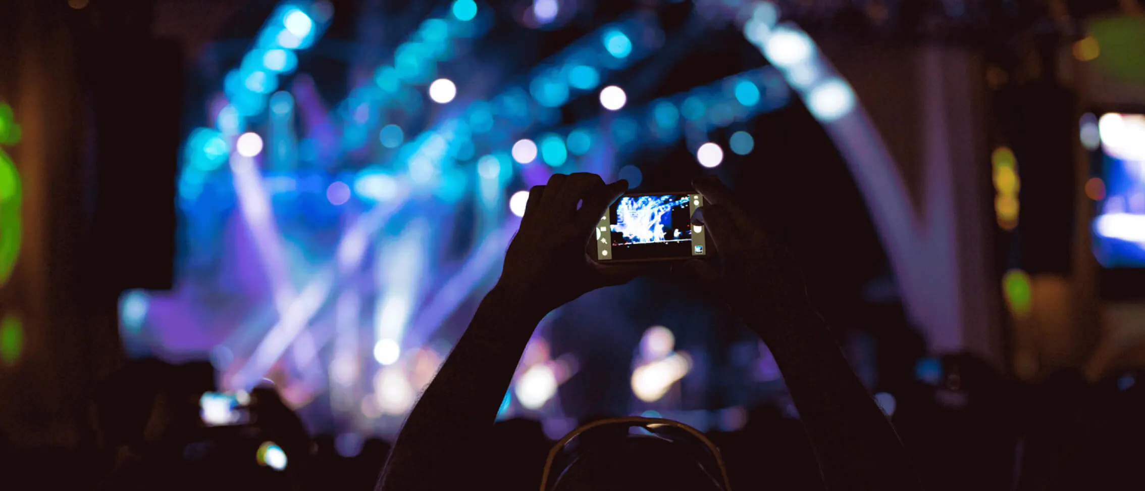 Guests capturing unforgettable moments on their phones as the concert lights up Universal Orlando Mardi Gras 2022 with vibrant blue and purple stage effects in the background.