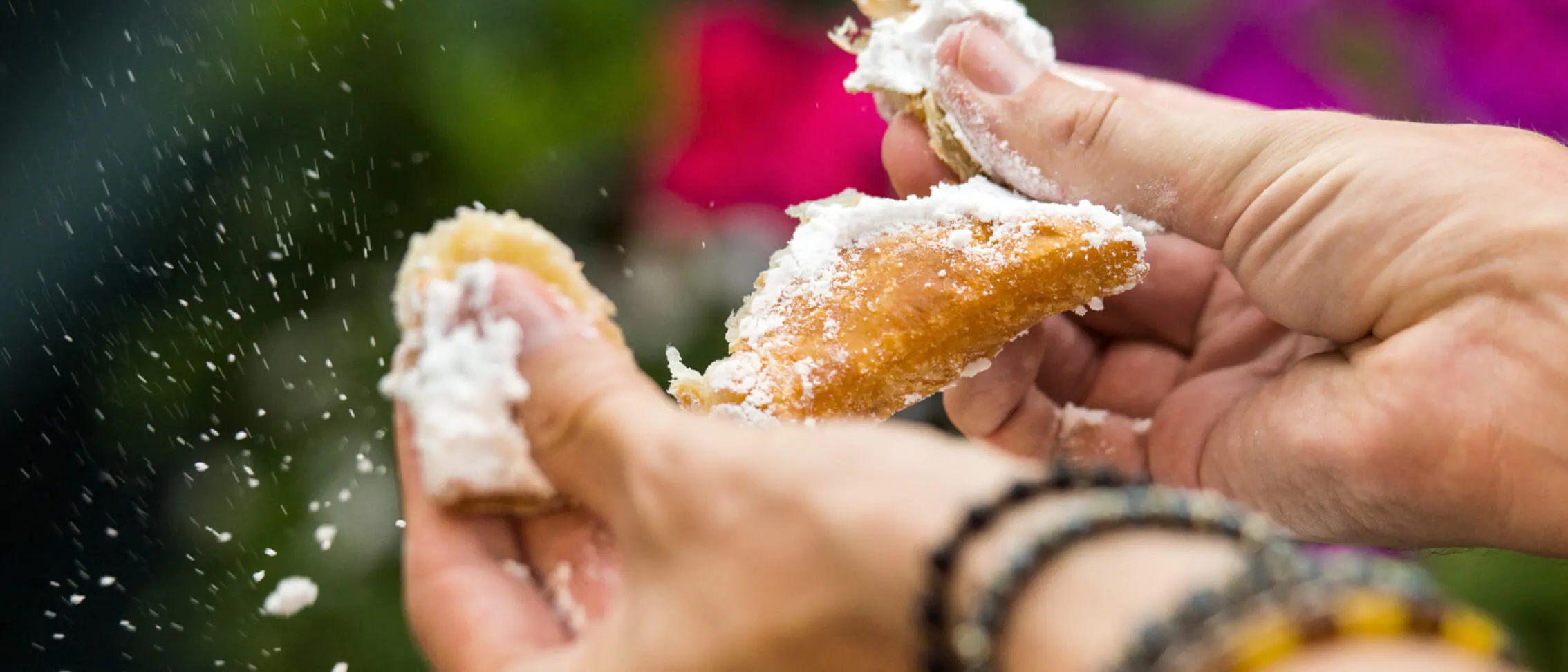 Powdered sugar cascades as hands split open a pastry, set against the vibrant blur of Universal Orlando’s Mardi Gras celebration in 2022, highlighting the festive and indulgent atmosphere that theme park guests enjoy.