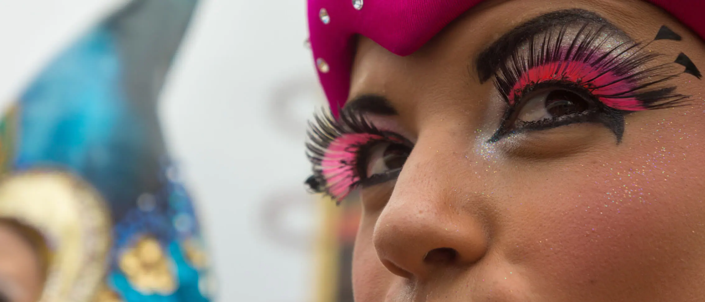 A dazzling Mardi Gras performer in bright makeup, dramatic false lashes, and a sparkling pink and blue jeweled costume adds vibrant energy to Universal Orlando’s 2022 Mardi Gras celebration.