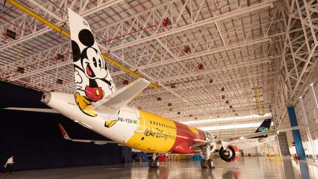 A vibrant Disney-themed plane featuring Mickey Mouse on its tail waits inside a spacious hangar, ready to whisk guests away for magical adventures above Brazil.