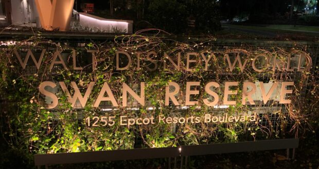 The Walt Disney World Swan Reserve sign glows brilliantly against the night sky, showcasing the resort’s newest addition with dazzling lights. Lush greenery and winding vines frame the entrance, immersing guests in that signature Disney magic as they arrive for their stay.