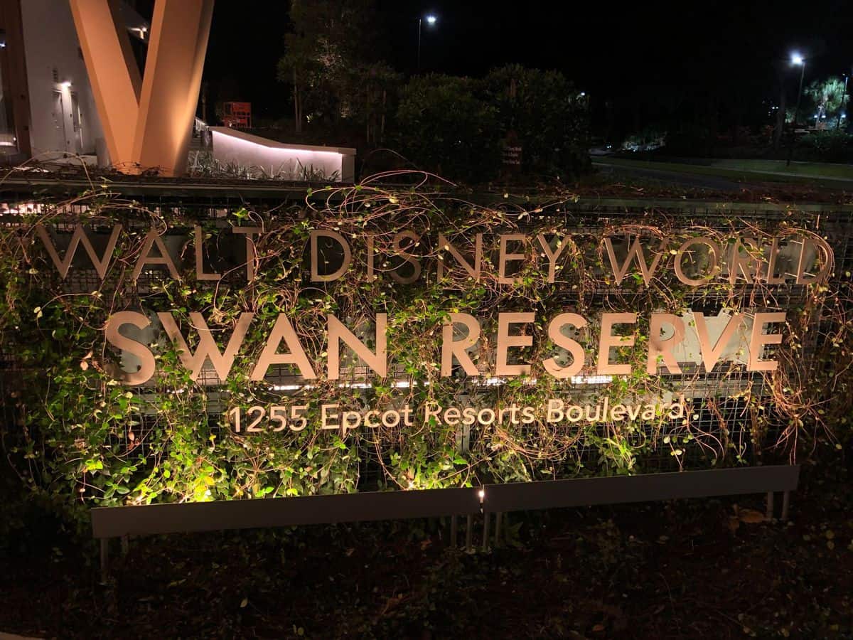 The Walt Disney World Swan Reserve sign glows brilliantly against the night sky, showcasing the resort’s newest addition with dazzling lights. Lush greenery and winding vines frame the entrance, immersing guests in that signature Disney magic as they arrive for their stay.