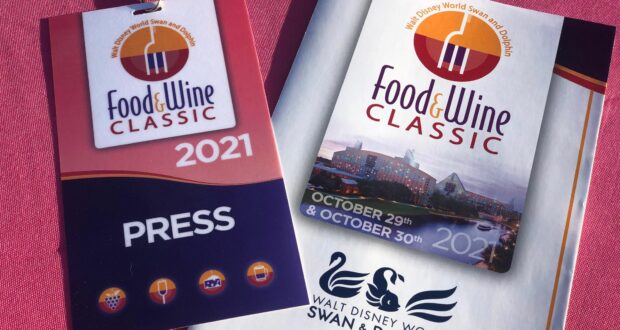 A Food & Wine Classic 2021 press badge and event flyer from the Walt Disney World Swan and Dolphin Resort displayed on a vibrant pink surface, capturing the excitement of Disney’s signature culinary event.