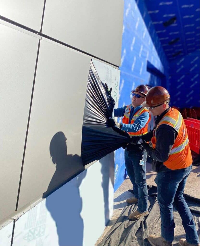 Two construction crew members carefully peel away a protective sheet from the facade of a new Disney attraction, their teamwork highlighting the behind-the-scenes precision that brings epic experiences like Guardians of the Galaxy: Cosmic Rewind to life at EPCOT. Sunlight glints off the panel as they work together to reveal another piece of Disney magic in progress.