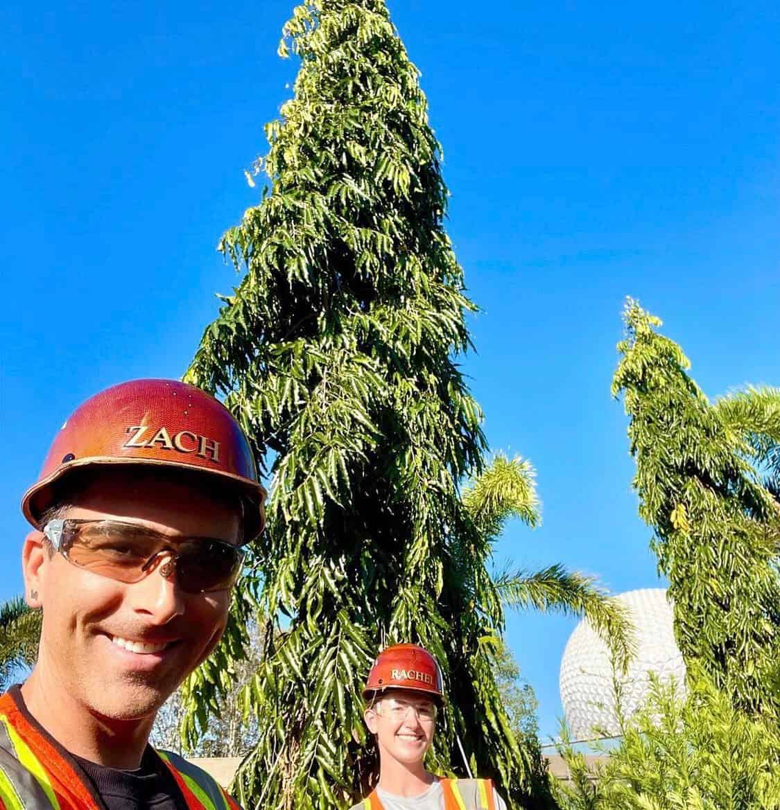 Cast Members in bright safety vests and helmets flash big smiles on Planting Day at EPCOT, standing outdoors among soaring trees with Spaceship Earth gleaming behind them.