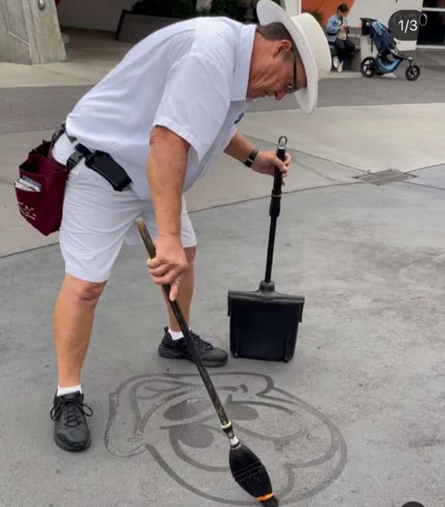 Mickey Mouse, dressed in classic white attire, entertains guests by creating delightful Water Art—a cheerful bear face—on the pavement with a wet broom, capturing the playful spirit of street performances at Walt Disney World.