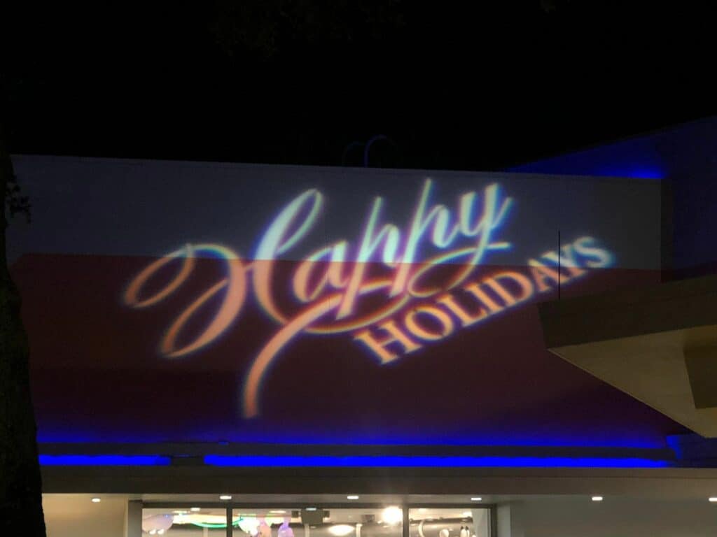 The phrase "Happy Holidays" shines in elegant script on a Disney park building at night, with blue accent lighting below setting a festive scene that captures the enchanting spirit of Very Merriest After Hours.