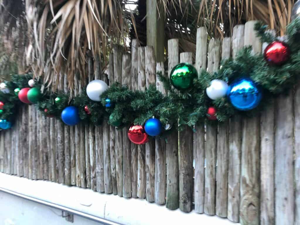 Festive Christmas garland adorned with vibrant ornaments decorates a wooden fence at Busch Gardens’ Christmas Town, with palm leaves above adding a tropical touch to the holiday scene.