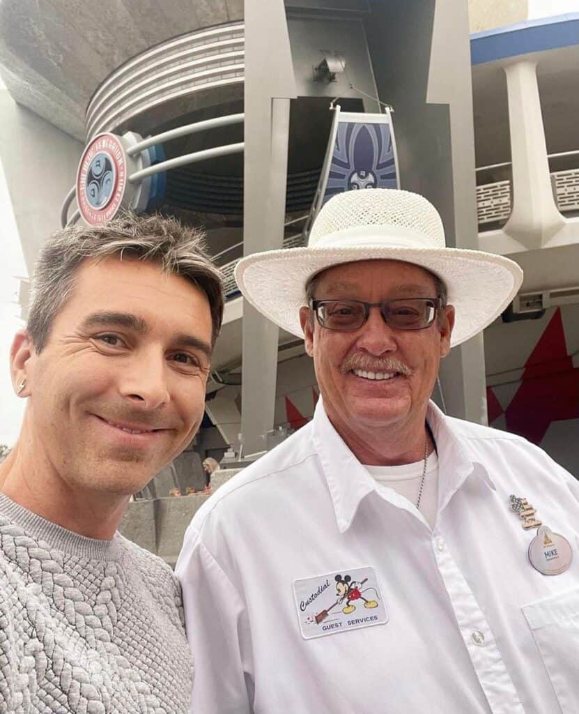Disney Cast Member in a themed guest services shirt and hat smiles alongside another guest in front of a strikingly futuristic Epcot pavilion, embodying the excitement and magic of Walt Disney World.