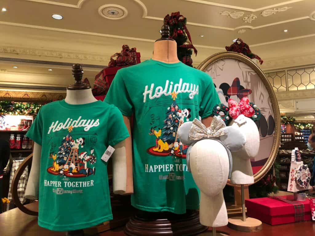 On display at the Disney Parks, two mannequins wear festive green "Holidays Happier Together" shirts alongside a pair of Minnie ear headbands, all part of the exclusive Holiday Merchandise collection inspired by Disney’s Very Merriest celebration.