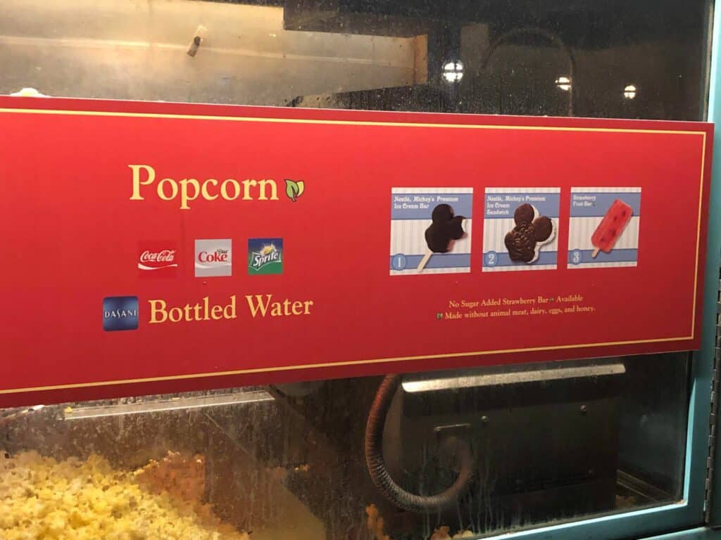 A bright red menu sign sits above buckets of popcorn, listing fan-favorite snacks like soda, bottled water, ice cream bars, and a classic strawberry popsicle—must-haves during Disney Very Merriest After Hours at Magic Kingdom. Surrounded by festive decor and old-school Disney treats below the sign, grabbing a snack here captures all the fun of a special after-hours celebration in the park.