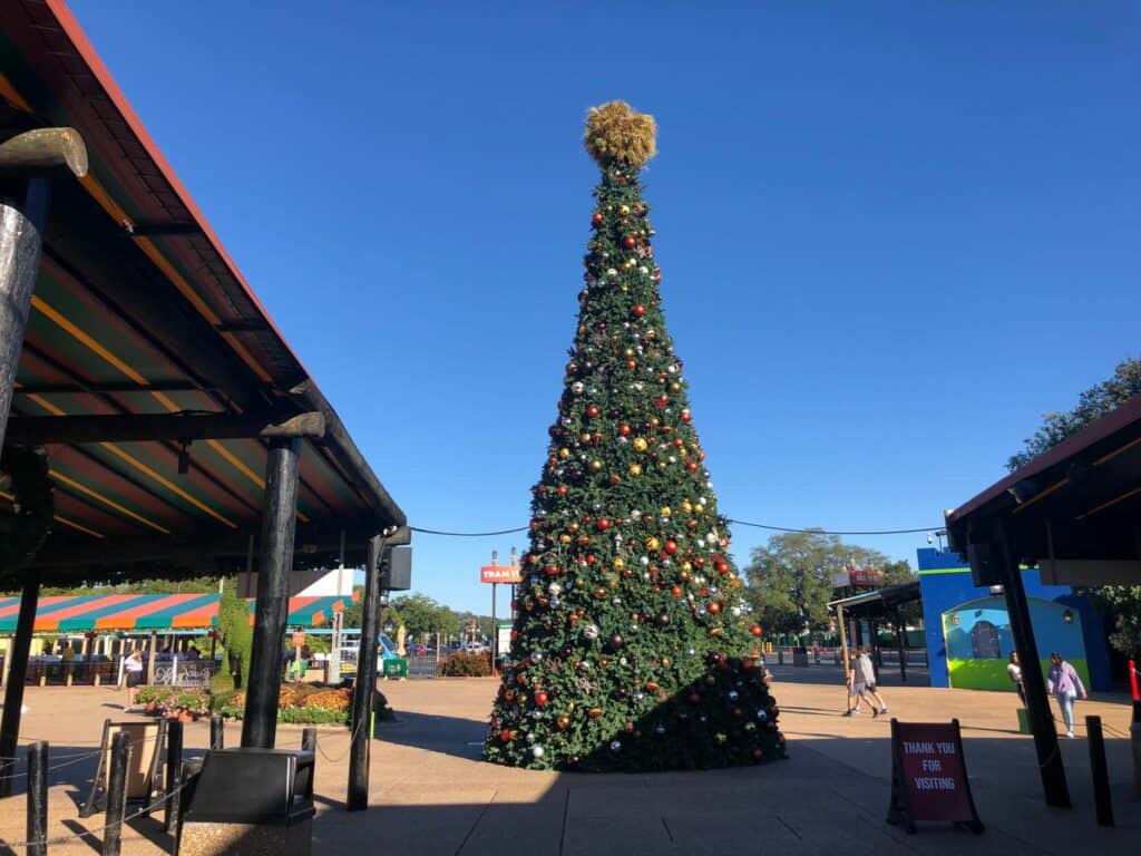 The grand Busch Gardens Christmas Town tree glimmers with festive decorations in the heart of the plaza, set against a brilliant blue sky.