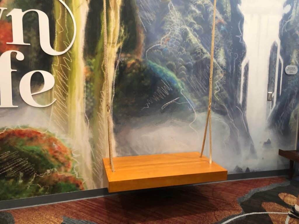 A whimsical wooden swing with thick rope is suspended indoors at Cirque du Soleil’s Drawn to Life, set before a vibrant mural of cascading waterfalls and dynamic, colorful shapes that capture the show’s imaginative staging and artistry.