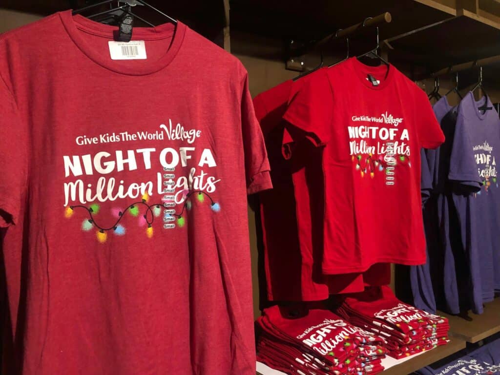Night of a Million Lights souvenir T-shirts in vibrant red are displayed hanging and folded on merch shelves, while nearby, bold purple Give Kids the World event shirts add extra excitement for collectors and fans at the attraction.