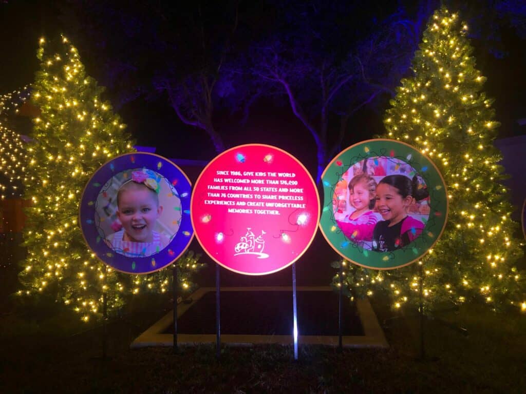 Illuminated signs featuring smiling children's faces and uplifting messages glow among festively decorated Christmas trees with sparkling lights, capturing the immersive holiday wonder of Give Kids The World's Night of a Million Lights event.