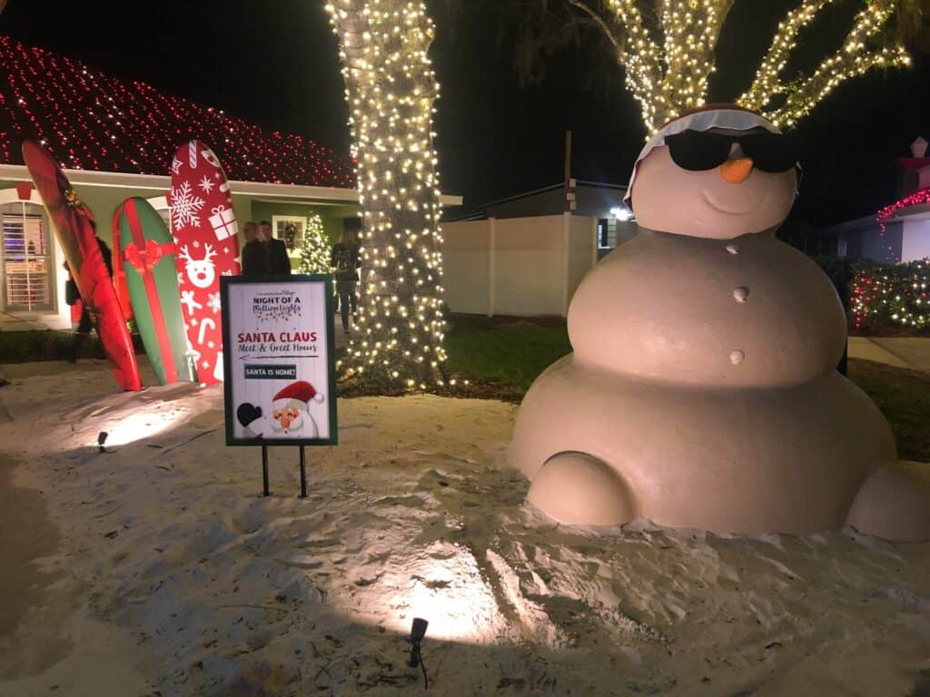 The iconic Give Kids the World snowman, rocking cool sunglasses, welcomes guests beside a festive Santa sign and beachy surfboards, all aglow with millions of holiday lights at Night of a Million Lights.