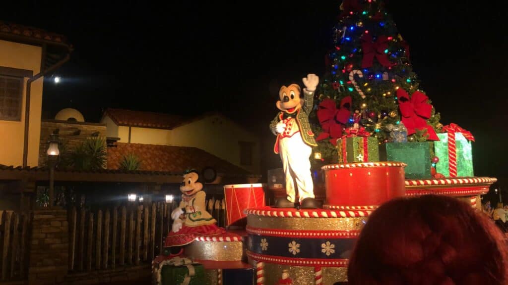 Mickey and Minnie Mouse, dressed in festive holiday outfits, wave to delighted guests from a dazzling Christmas float with a beautifully decorated tree, lighting up the night during Disney Very Merriest After Hours at Magic Kingdom—the perfect way to experience Disney magic during the holidays.