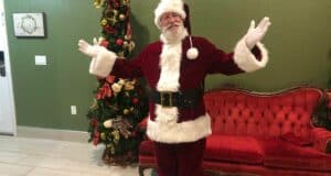 Santa Claus at our holiday attraction welcomes guests with open arms, standing by a festive red couch and a sparkling Christmas tree. He greets families who’ve reserved their Meet Santa experience, enjoying exclusive access without the long lines thanks to our Virtual Queue system.
