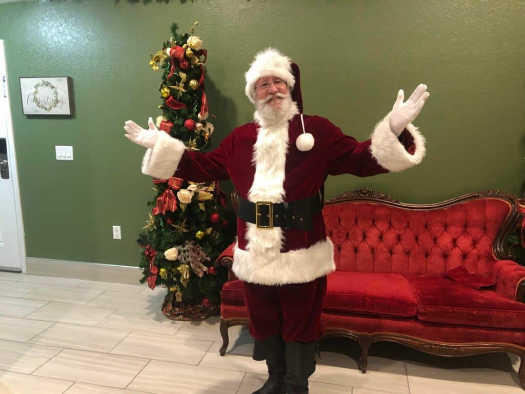 Santa Claus welcomes guests with a cheerful smile and open arms beside a festive red couch and sparkling Christmas tree inside Give Kids the World Village, ready to create holiday memories during the magical Night of a Million Lights event.