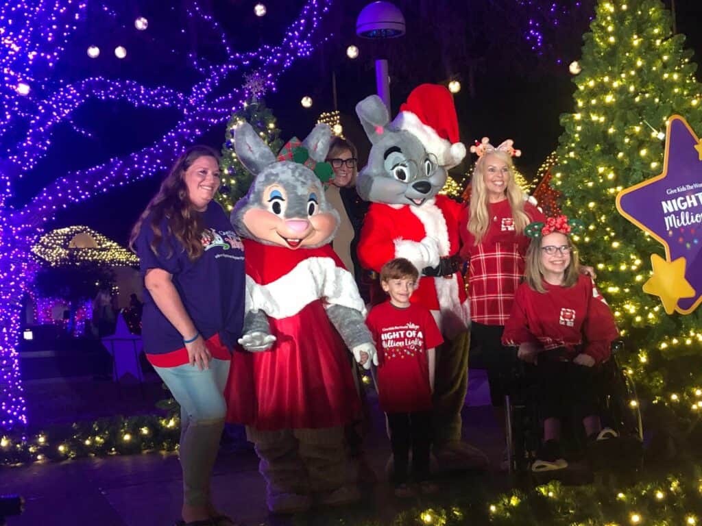 Guests enjoying Night of a Million Lights at Give Kids the World, posing with two iconic Village bunnies in costume, surrounded by festive Christmas trees and vibrant purple holiday lights.