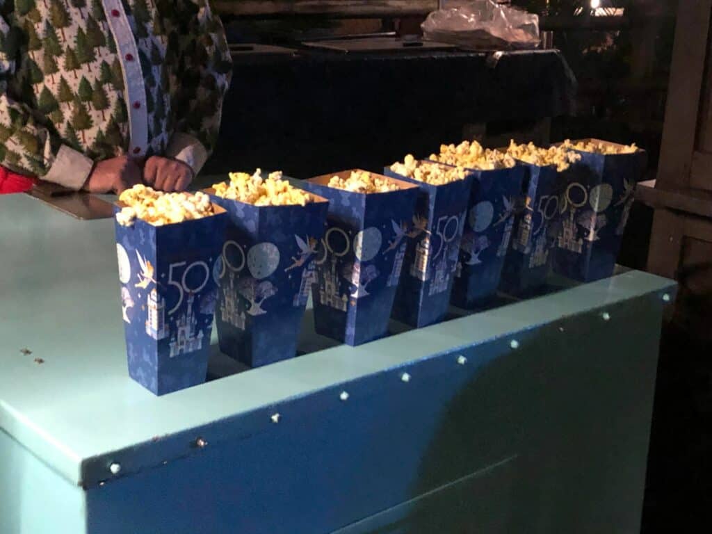 Seven blue popcorn boxes lined up on a teal counter at Disney's Very Merriest After Hours, with a guest in a festive tree-patterned sweater eagerly ready to enjoy the holiday treats.