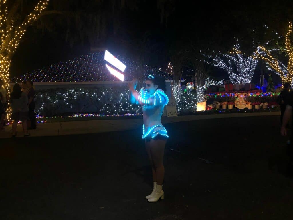 A glowing blue costumed entertainer takes center stage on a festively illuminated street, surrounded by sparkling holiday trees during the Night of Million Lights celebration at Give Kids the World Village.