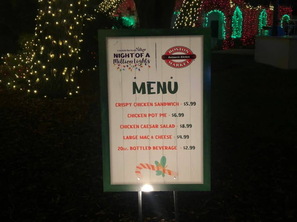 Festive menu board illuminated with sparkling holiday lights at Give Kids the World’s Night of a Million Lights event, featuring theme park favorites like chicken sandwiches, pot pies, Caesar salad, mac & cheese, and assorted drinks.