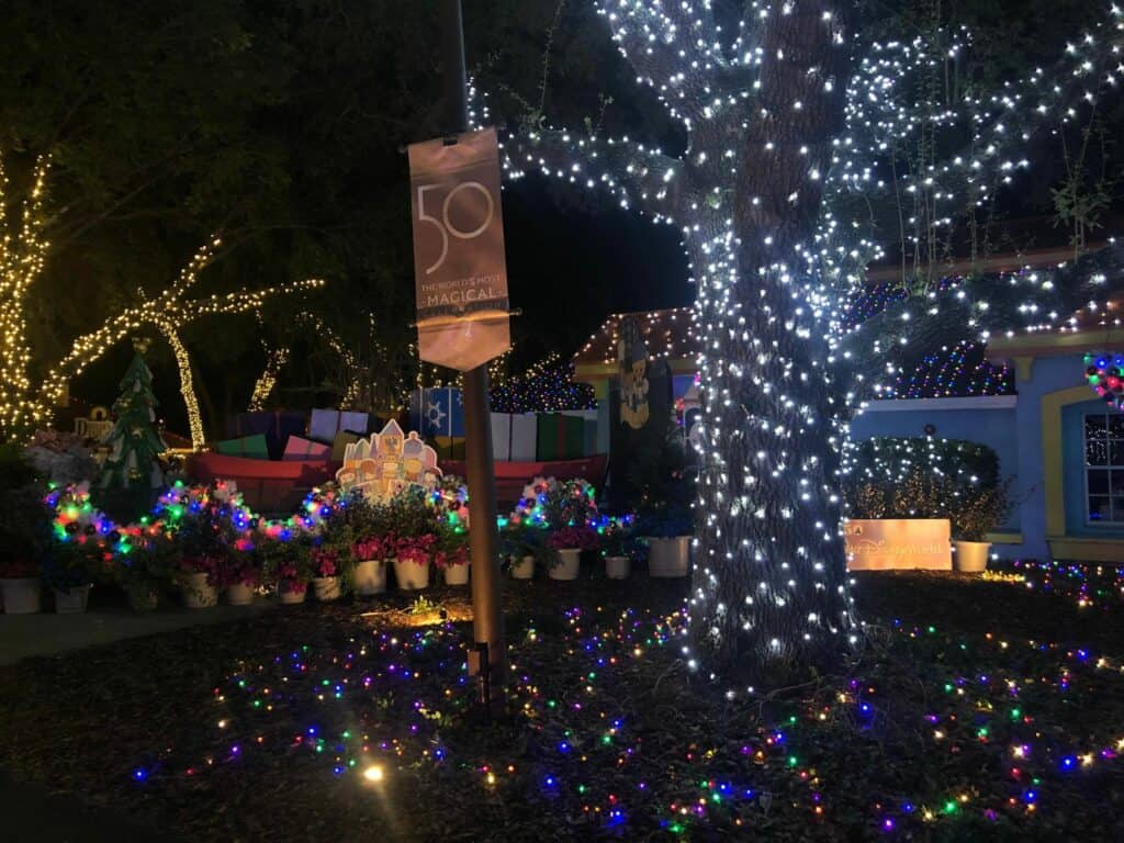 A festive scene at Give Kids the World’s Night of a Million Lights, featuring a house and towering tree dazzling with vibrant holiday lights, highlighted by a sparkling “50 Years Magical” banner celebrating the legacy of Walt Disney World Resort.