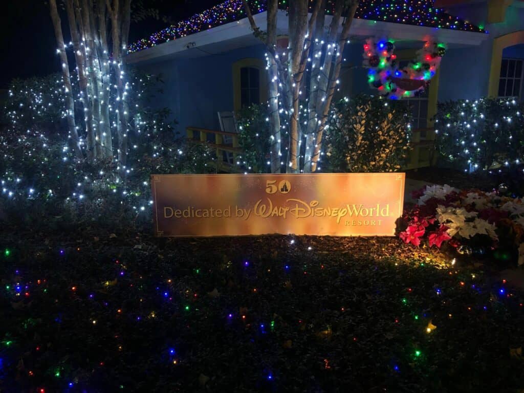 A magical nighttime display at Give Kids the World's Night of a Million Lights, featuring Disney World-style trees twinkling with festive lights, a whimsical house, and a sign proudly reading "Dedicated by Walt Disney World Resort.