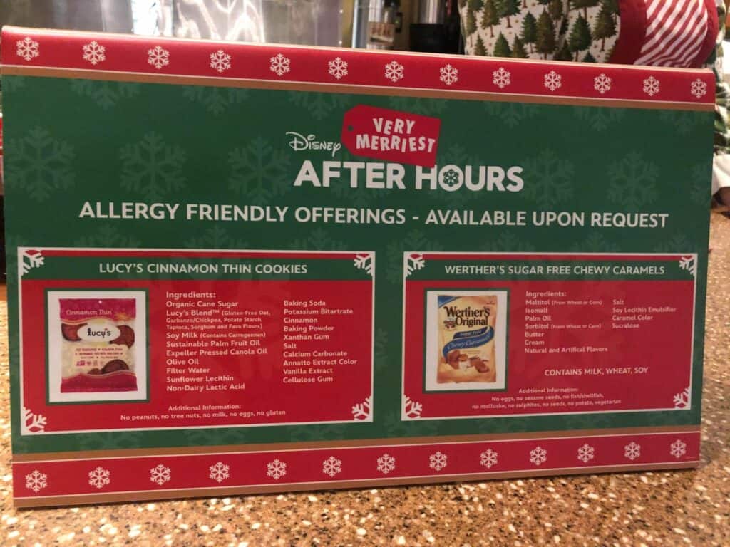 Disney Very Merriest sign showcases allergy-friendly cookies and caramel candies with clear ingredient lists on the countertop, inviting fans to grab exclusive After Hours treats while checking out Holiday Merch in the festive park setting.
