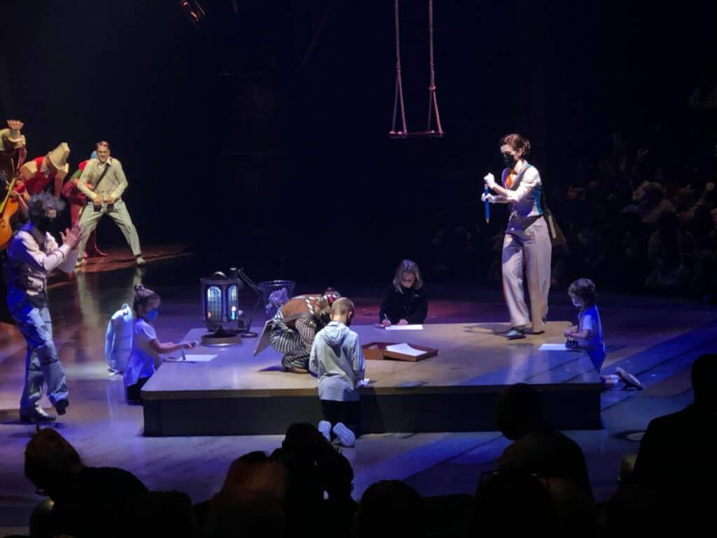 On the Cirque du Soleil Drawn to Life stage, families and fans watch as children and adults sketch together on the floor beneath theatrical, moody lighting, capturing the signature whimsical flair of opening night at Walt Disney World’s Disney Springs.