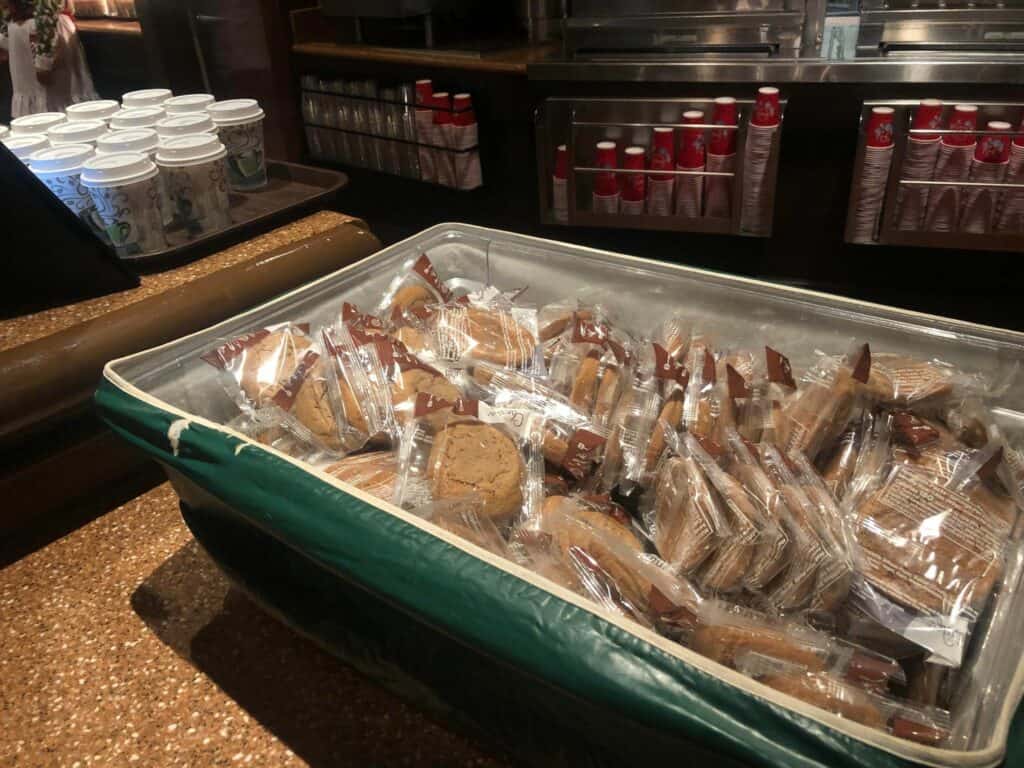 A festive selection of individually wrapped cookies waits in a bin, while stacks of coffee cups and lids line the counter behind—an ideal treat to fuel your Disney Very Merriest After Hours adventure or to enjoy alongside this year’s exclusive Holiday Merchandise.
