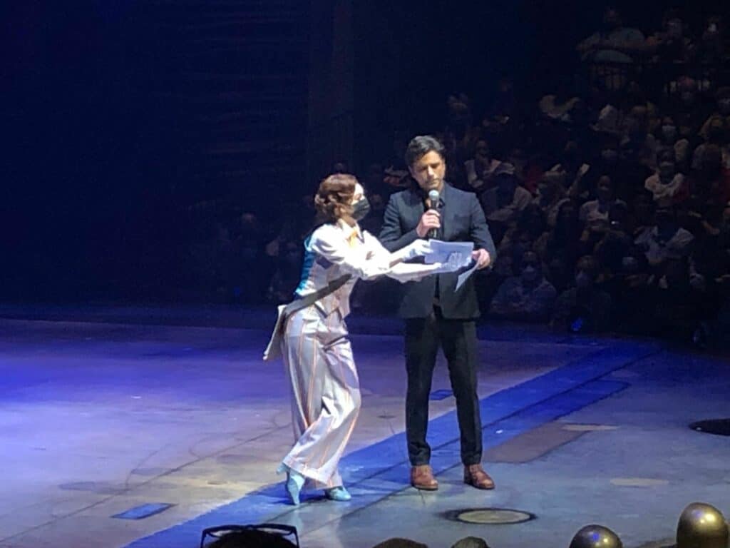 Cirque du Soleil’s Opening Night electrifies the arena as a woman in a white suit and a man in a dark suit address the buzzing crowd from center stage, igniting anticipation for an unforgettable entertainment experience.
